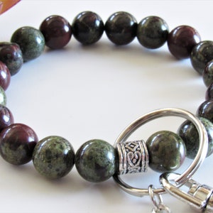 May include: A green and red gemstone beaded bracelet with a silver key ring and clasp. The bracelet is made of round beads and has a decorative silver bead with a design.