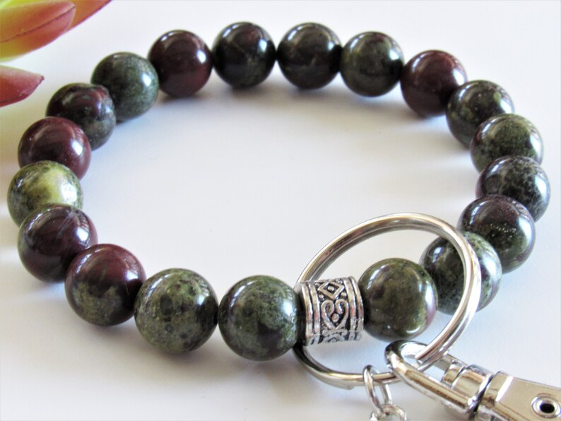 May include: A green and red gemstone beaded bracelet with a silver key ring and clasp. The bracelet is made of round beads and has a decorative silver bead with a design.