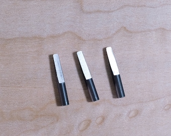 May include: Three silver and black metal screwdriver bits on a light wood surface.