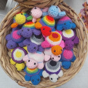 Könnte beinhalten: Ein Weidenkorb voller bunter, gehäkelter Schildkrötenspielzeuge. Die Schildkröten haben verschiedene Farben, darunter Blau, Lila, Rosa, Gelb und Orange, mit Regenbogenakzenten. Jede Schildkröte hat kleine schwarze Augen und ist aus weichem Garn gefertigt.