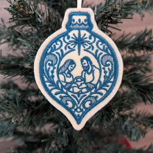 May include: A cream-coloured felt ornament shaped like a Christmas bauble, featuring intricate blue embroidery of a nativity scene with a star above. The design includes decorative filigree patterns surrounding the figures and a white hanging ribbon.