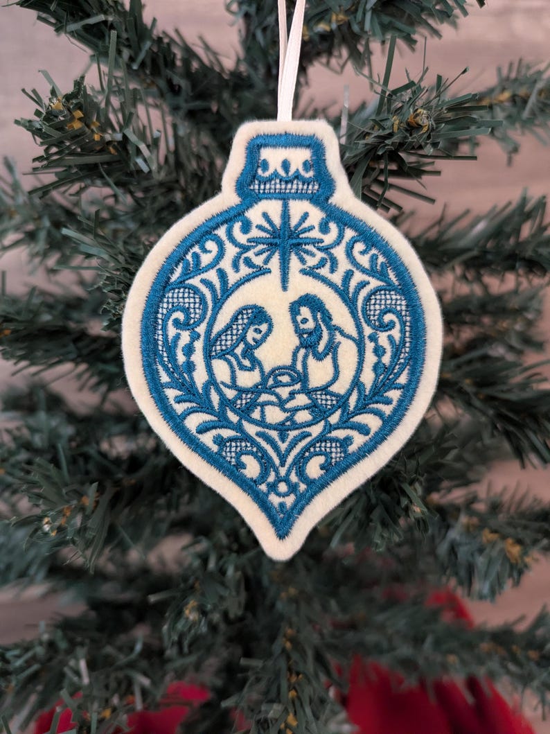 May include: A cream-coloured felt ornament shaped like a Christmas bauble, featuring intricate blue embroidery of a nativity scene with a star above. The design includes decorative filigree patterns surrounding the figures and a white hanging ribbon.
