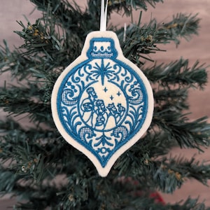May include: A teardrop-shaped felt Christmas ornament featuring intricate blue embroidery. The design depicts three figures in a central circular frame, surrounded by ornate scrollwork and a star motif at the top, suspended by a white ribbon.