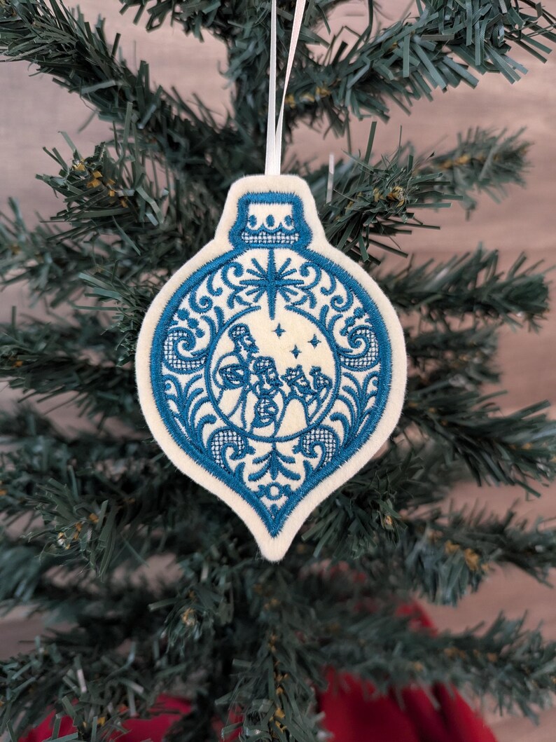 May include: A teardrop-shaped felt Christmas ornament featuring intricate blue embroidery. The design depicts three figures in a central circular frame, surrounded by ornate scrollwork and a star motif at the top, suspended by a white ribbon.