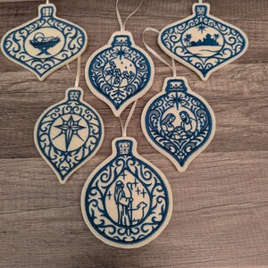 May include: Six white felt Christmas ornaments with blue embroidery featuring nativity scenes, including a manger, the star of Bethlehem, the three wise men, a shepherd with a sheep, and a holy family, each with a white ribbon loop for hanging.