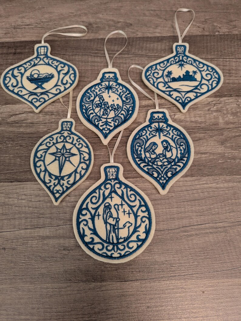 May include: Six white felt Christmas ornaments with blue embroidery featuring nativity scenes, including a manger, the star of Bethlehem, the three wise men, a shepherd with a sheep, and a holy family, each with a white ribbon loop for hanging.