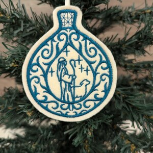 May include: A cream-coloured felt ornament shaped like a bottle, featuring blue machine embroidery of a shepherd with a staff and a sheep. The design includes intricate blue scrollwork patterns surrounding the central figure and stars.