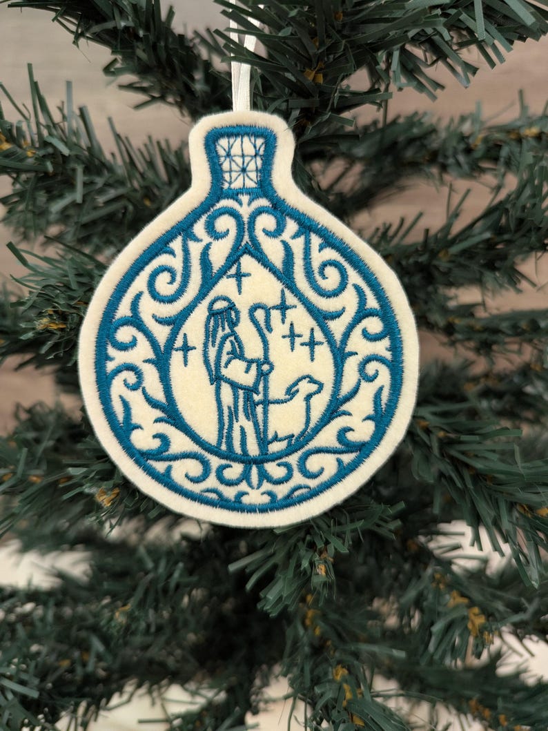 May include: A cream-coloured felt ornament shaped like a bottle, featuring blue machine embroidery of a shepherd with a staff and a sheep. The design includes intricate blue scrollwork patterns surrounding the central figure and stars.