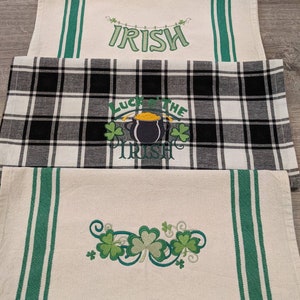 May include: Three St. Patrick's Day themed table runners. One is cream-colored with green stripes and the word "IRISH". Another is black and white plaid with "Luck of the Irish". The last is cream-colored with green shamrocks.