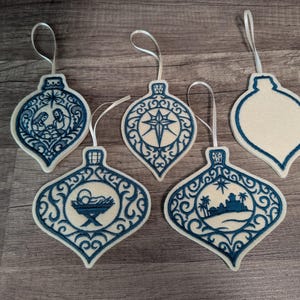 May include: Five teardrop-shaped ornaments made of off-white felt with dark blue embroidery. Four ornaments feature intricate filigree borders and central designs including a nativity scene, a star, a manger, and a desert landscape. One is blank.