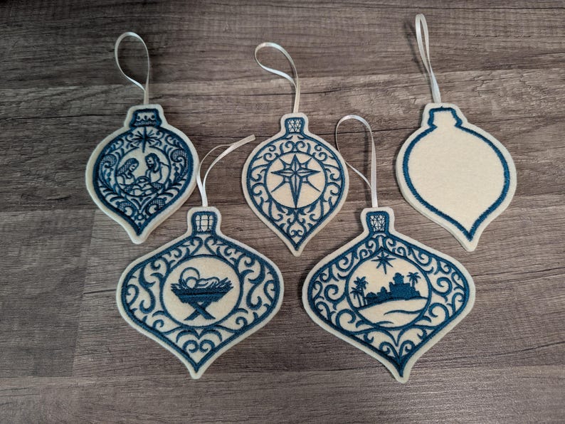 May include: Five teardrop-shaped ornaments made of off-white felt with dark blue embroidery. Four ornaments feature intricate filigree borders and central designs including a nativity scene, a star, a manger, and a desert landscape. One is blank.