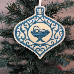 May include: A cream-coloured felt Christmas ornament shaped like a bauble, featuring blue machine embroidery. The design includes an intricate scrollwork border and a central depiction of a baby in a manger. A white ribbon loop is attached.