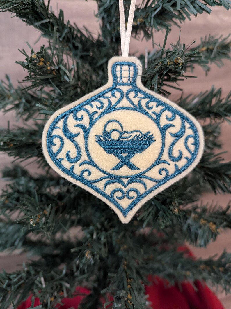 May include: A cream-coloured felt Christmas ornament shaped like a bauble, featuring blue machine embroidery. The design includes an intricate scrollwork border and a central depiction of a baby in a manger. A white ribbon loop is attached.