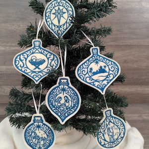 May include: Five teardrop-shaped Christmas ornaments featuring intricate blue embroidery on a cream-coloured felt background. Each ornament displays a unique religious scene, including a star, a manger, a desert landscape, a nativity, and a shepherd.