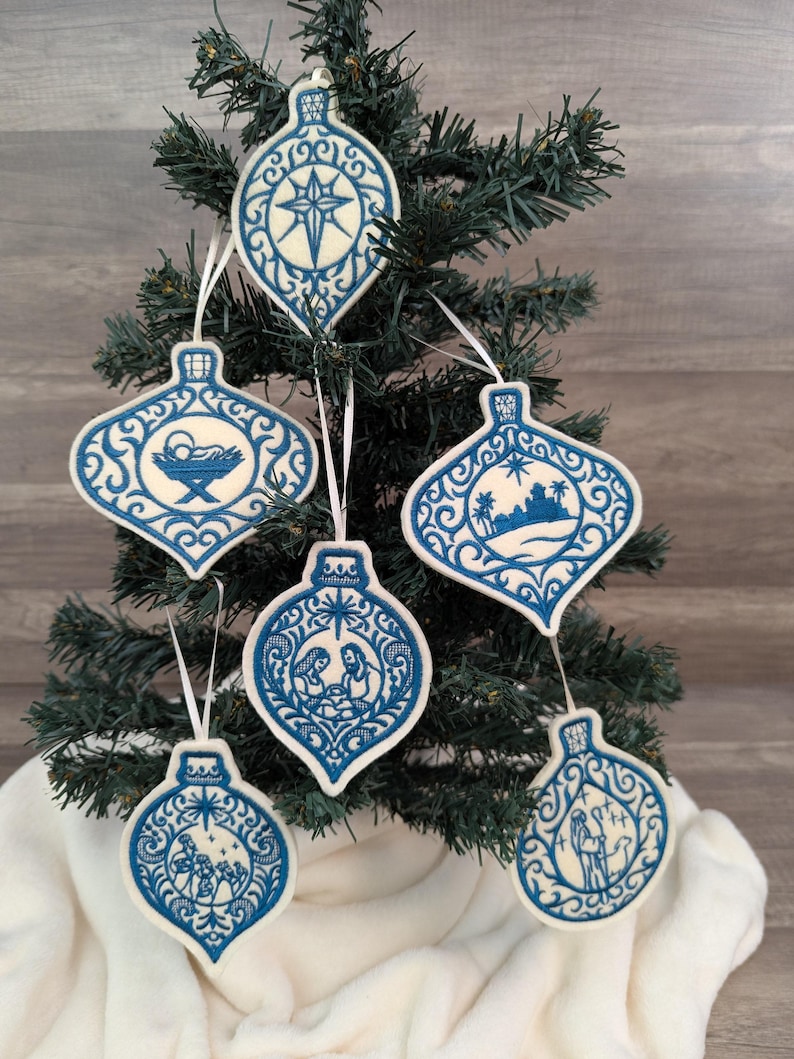May include: Five teardrop-shaped Christmas ornaments featuring intricate blue embroidery on a cream-coloured felt background. Each ornament displays a unique religious scene, including a star, a manger, a desert landscape, a nativity, and a shepherd.