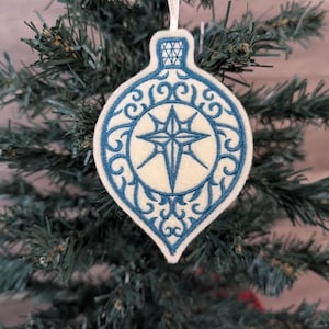 May include: A teardrop-shaped felt Christmas ornament featuring intricate blue embroidery. The design includes a central star motif surrounded by swirling filigree patterns and a geometric cap detail. A thin white ribbon is attached for hanging.