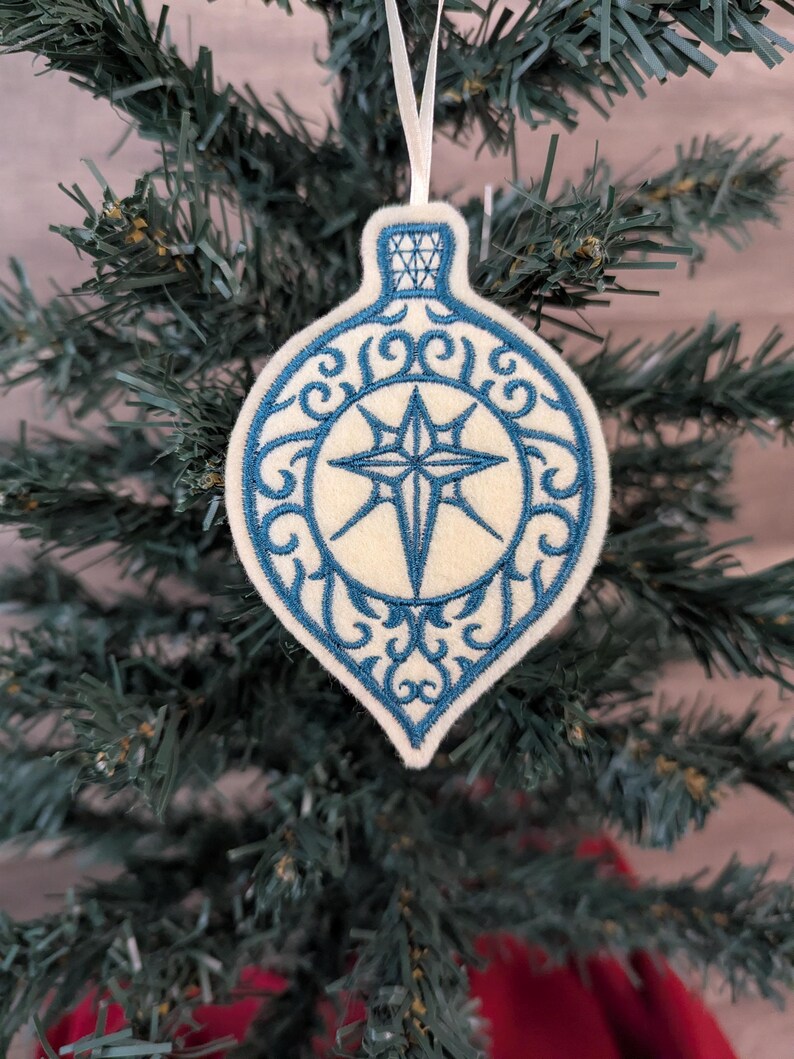 May include: A teardrop-shaped felt Christmas ornament featuring intricate blue embroidery. The design includes a central star motif surrounded by swirling filigree patterns and a geometric cap detail. A thin white ribbon is attached for hanging.