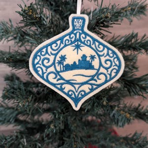 May include: A cream-coloured felt ornament shaped like a teardrop, featuring intricate blue embroidery of a desert landscape with palm trees and a star above. The design is framed by ornate blue scrollwork and hangs from a white ribbon loop.