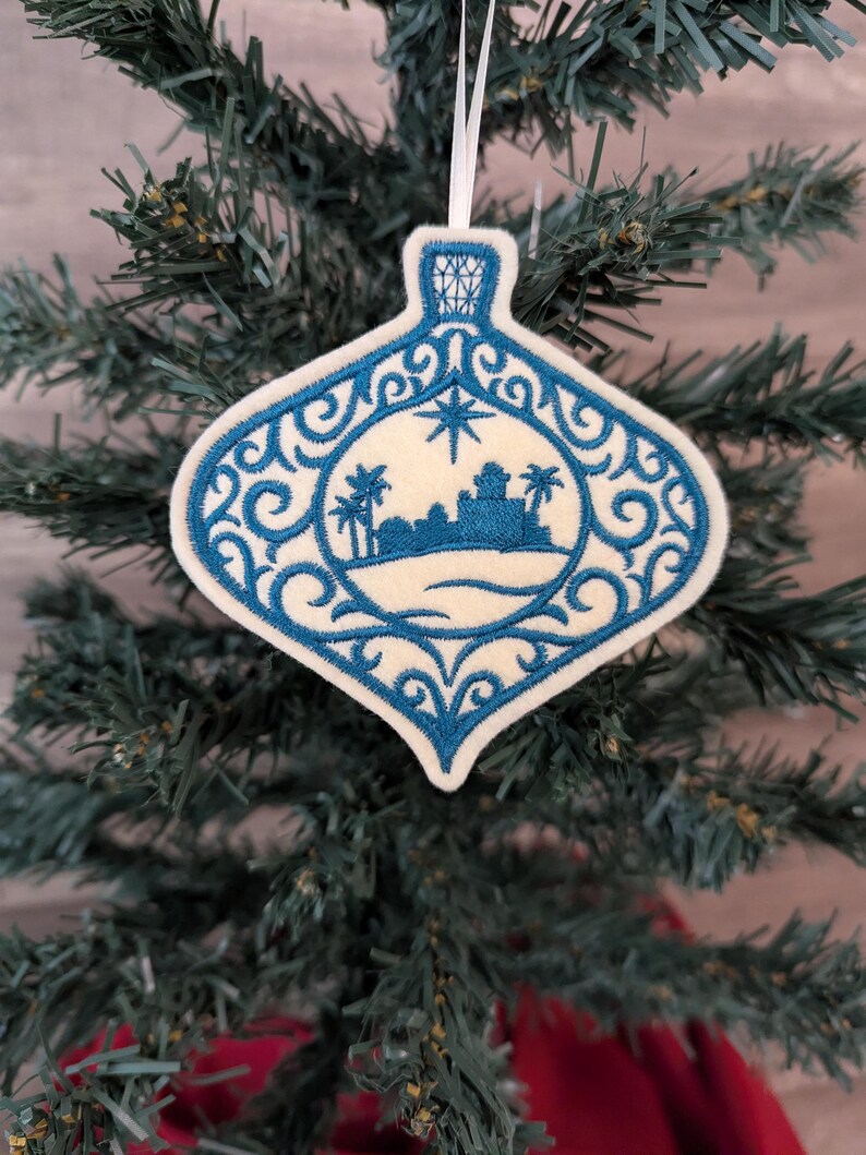 May include: A cream-coloured felt ornament shaped like a teardrop, featuring intricate blue embroidery of a desert landscape with palm trees and a star above. The design is framed by ornate blue scrollwork and hangs from a white ribbon loop.