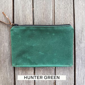 May include: A rectangular hunter green zippered pouch featuring a black zipper closure and a thin brown leather pull tab. The pouch is constructed from a textured fabric and includes the text HUNTER GREEN printed in black on a white rectangular label.