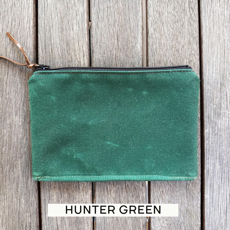May include: A rectangular hunter green zippered pouch featuring a black zipper closure and a thin brown leather pull tab. The pouch is constructed from a textured fabric and includes the text HUNTER GREEN printed in black on a white rectangular label.