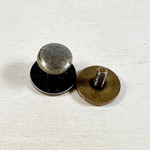 May include: A close-up of two antique brass-toned snap fasteners. The top fastener has a round head and a black base. The bottom fastener has a round base with a screw protruding from the center.