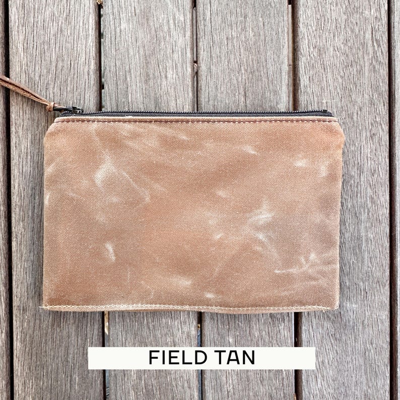 May include: A rectangular zippered pouch in a field tan color with a weathered, textured finish. The pouch features a black zipper closure along the top edge and a thin brown leather pull tab attached to the metal zipper slider for easy opening.