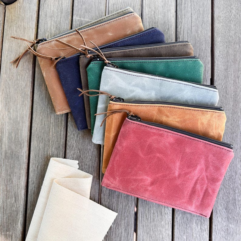 May include: A collection of rectangular zippered pouches made from waxed canvas in various colors including tan, navy, dark brown, forest green, light gray, mustard yellow, and deep red, each featuring a black zipper and a thin leather pull tab.