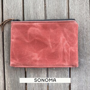 May include: A rectangular pouch made of a textured, rust-colored fabric with a black zipper closure along the top edge. A thin brown leather pull is attached to the zipper slider. The word SONOMA is printed in black text below the pouch.