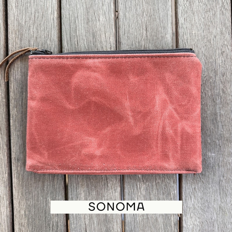 May include: A rectangular pouch made of a textured, rust-colored fabric with a black zipper closure along the top edge. A thin brown leather pull is attached to the zipper slider. The word SONOMA is printed in black text below the pouch.