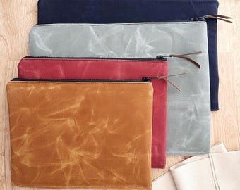 May include: A stack of four rectangular zippered pouches made of waxed canvas in navy blue, light gray, deep red, and tan. Each pouch features a black zipper closure with a thin brown leather pull tab attached to the metal slider for easy access.