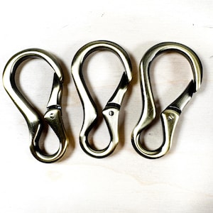 May include: Three antique brass-colored spring snap hooks. Each hook has a curved top, a hinged lever, and a spring-loaded closure. The hooks are arranged on a light-colored wooden surface.