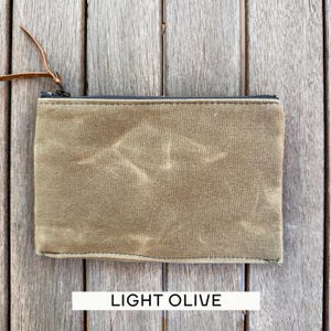 May include: A rectangular light olive zippered pouch made of a textured, waxed-style fabric. The pouch features a black zipper closure along the top edge and a thin, brown leather pull tab attached to the metal zipper slider for easy opening.