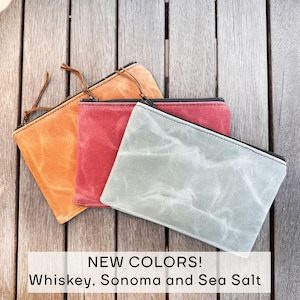 May include: Three rectangular zippered pouches in whiskey orange, Sonoma red, and sea salt gray. Each pouch features a textured, waxed canvas material with a black zipper closure and a thin leather pull tab. Text reads: NEW COLORS! Whiskey, Sonoma and Sea Salt.