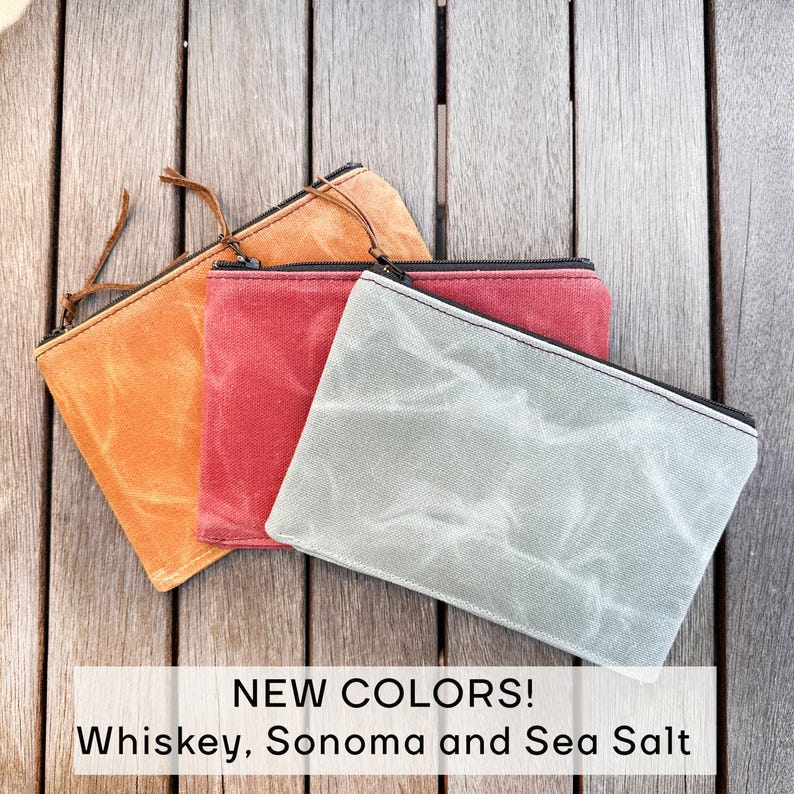 May include: Three rectangular zippered pouches in whiskey orange, Sonoma red, and sea salt gray. Each pouch features a textured, waxed canvas material with a black zipper closure and a thin leather pull tab. Text reads: NEW COLORS! Whiskey, Sonoma and Sea Salt.