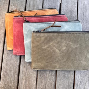 May include: Four rectangular zippered pouches made of waxed canvas in shades of mustard yellow, deep red, light sage green, and olive green. Each pouch features a black zipper closure with a thin brown leather pull tab attached to the slider.