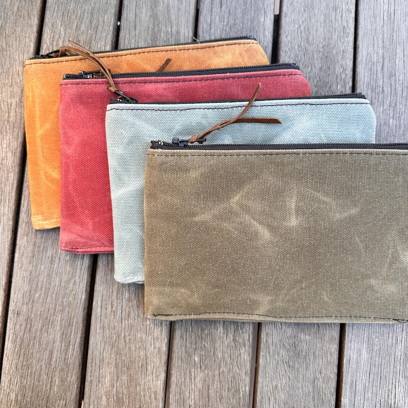 May include: Four rectangular zippered pouches made of waxed canvas in shades of mustard yellow, deep red, light sage green, and olive green. Each pouch features a black zipper closure with a thin brown leather pull tab attached to the slider.