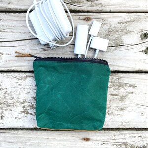 May include: A green waxed canvas zippered pouch with a brown leather zipper pull. The pouch is sitting on a white wooden surface with white cords and white adapters.