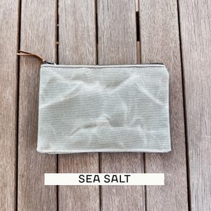 May include: A rectangular zippered pouch in a light sage green color with a textured, wrinkled fabric surface. The pouch features a dark metal zipper closure with a thin brown leather pull tab. The text SEA SALT is printed below the pouch.