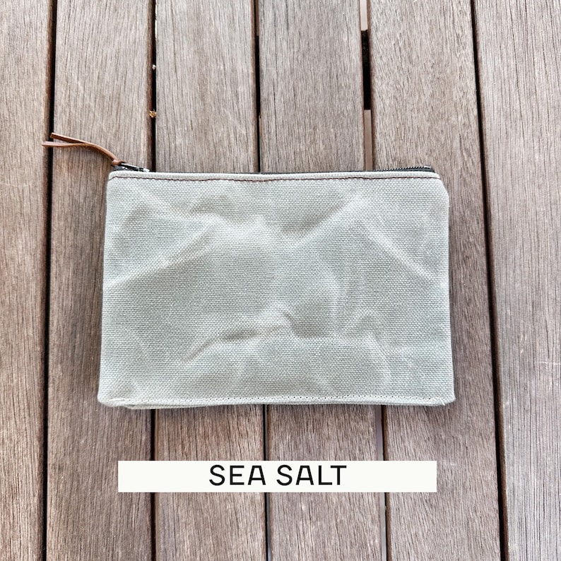 May include: A rectangular zippered pouch in a light sage green color with a textured, wrinkled fabric surface. The pouch features a dark metal zipper closure with a thin brown leather pull tab. The text SEA SALT is printed below the pouch.