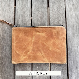 May include: A rectangular zippered pouch made of tan waxed canvas with a visible creased texture. The pouch features a black zipper closure along the top edge with a thin brown leather pull tab attached to the metal slider. The word WHISKEY is printed.