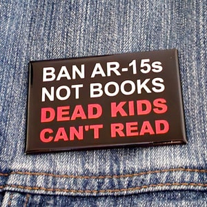 May include: A black and white button with the text "BAN AR-15s NOT BOOKS DEAD KIDS CAN'T READ" in white letters with the words "DEAD KIDS" in red letters. The button is pinned to a blue denim jacket.
