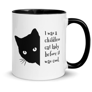 May include: White ceramic mug with a black rim and handle. The mug features a black silhouette of a cat's head peeking out from the side, with the text "I was a childless cat lady before it was cool." printed on the front.
