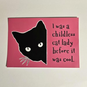 May include: A pink background with a black cat peeking out from the right side. The text reads "I was a childless cat lady before it was cool."