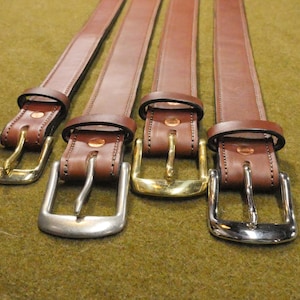May include: Four brown leather belts with different metal buckle styles: gold, silver, and brass. The belts are arranged side by side, showcasing the variety of hardware options. The leather appears to be of high quality.