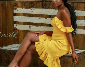 May include: A woman wearing a yellow off-the-shoulder dress with ruffles sits on a white and black wooden bench with an iron frame. The bench is in front of a wooden wall and a window with a view of green foliage.
