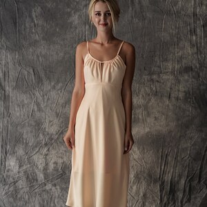 May include: A peach-colored midi dress with a scoop neck and thin straps. The dress has a fitted bodice and a flowing skirt. The model is wearing black heeled sandals.
