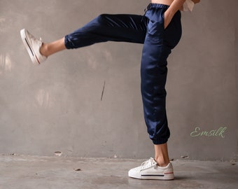 Puede incluir: Una mujer con un top blanco de tirantes y pantalones de satén azul oscuro. Está de pie con una pierna levantada y lleva zapatillas blancas. El fondo es una pared gris con el texto "Emsilk" en verde.