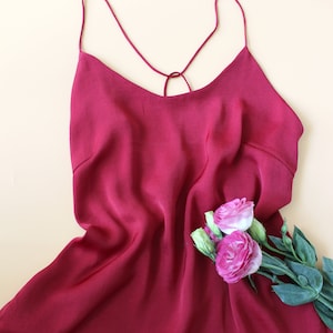 Puede incluir: Un top de satén burdeos con tirantes finos y un detalle de lazada en la espalda. El top está colocado sobre una superficie beige claro con un ramo de flores rosas.