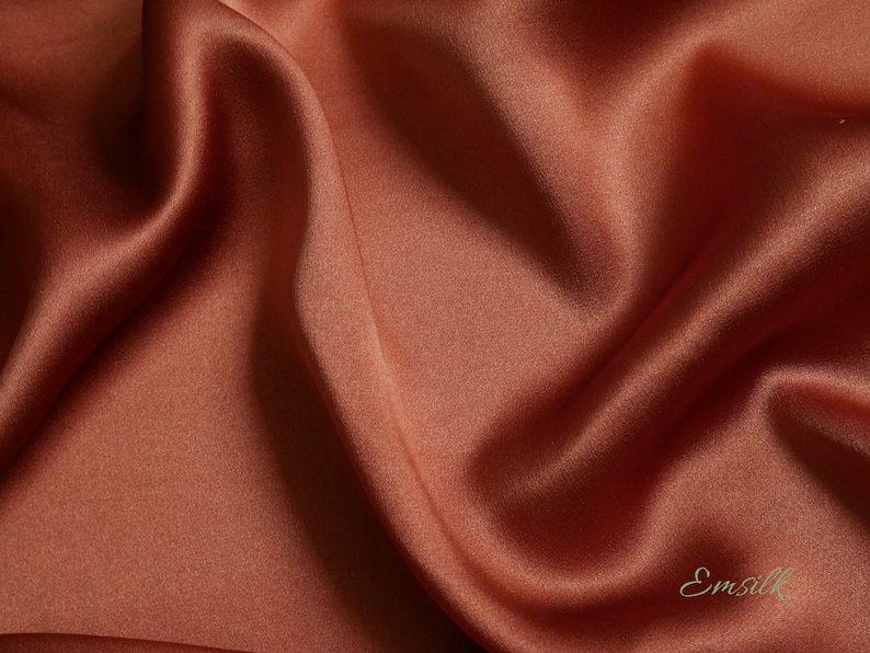 May include: Close-up of a flowing, rust-colored silk fabric. The fabric has a smooth, lustrous texture with soft folds and highlights. The word "Emsilk" is printed in a cursive font in the bottom right corner.